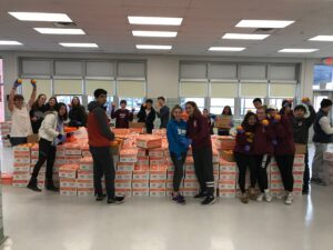 Group of volunteers standing behind stacks of boxed supplies in a large indoor room.