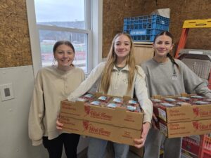 Three women smiling, holding boxes of food items indoors.