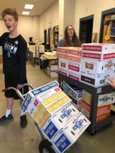 Two kids with stacked boxes and a cart in a room.