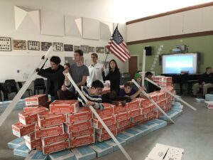 Group posing on a makeshift raft made of pizza boxes indoors.