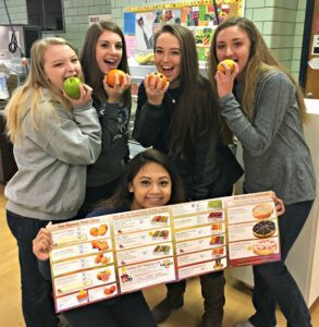 Four young women holding apples and a menu, smiling playfully.
