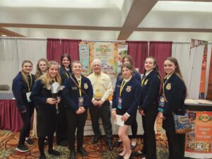 A group of young women in formal attire posing indoors with a man in a yellow shirt.