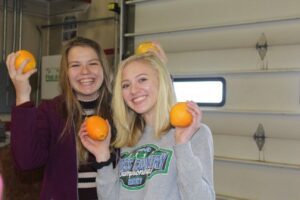 Two young women smiling and holding oranges indoors.